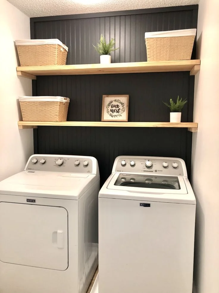 Small laundry closet featuring top-load Maytag washer and dryer, dark black vertical beadboard accent wall, natural wood floating shelves with woven storage baskets, and green potted plants.