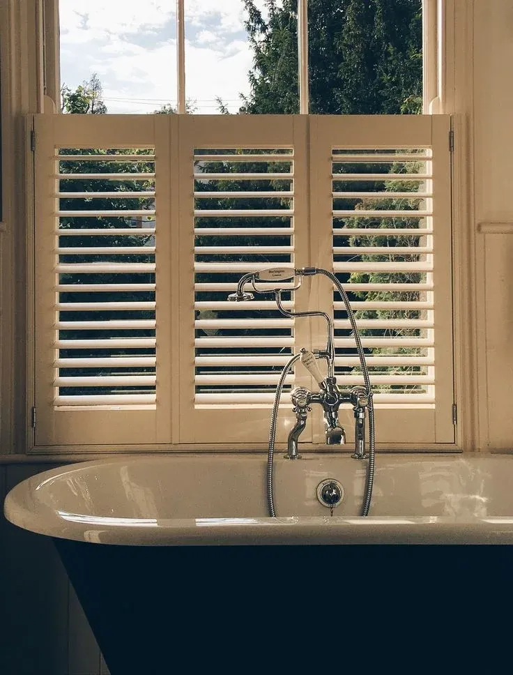 A vintage clawfoot bathtub set below a multi-pane window featuring white cafe-style plantation shutters on the bottom half for bathroom privacy.