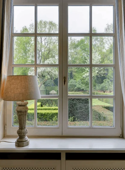 A tall white wooden grid window framed by neutral drapes, with a rustic table lamp on the window sill looking out onto a manicured green garden.