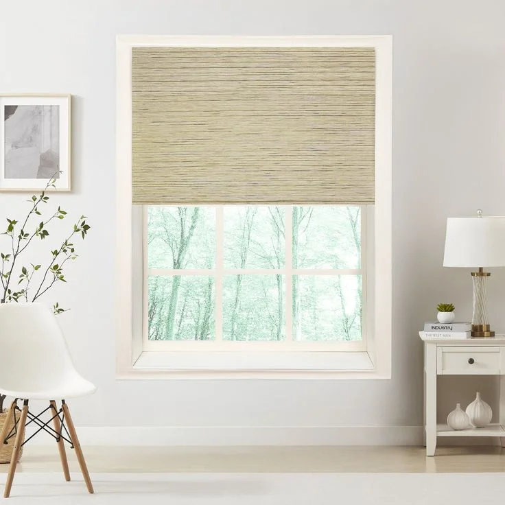 A light gray-green woven bamboo roller shade is partially lowered over a bright window with multiple panes, looking out onto a treed view. It is styled in a modern room with a white chair and white side table.