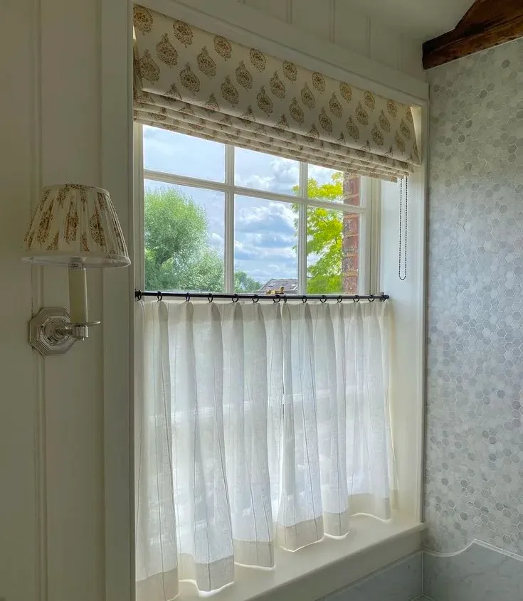 White grid window featuring a beige patterned Roman blind and a sheer white cafe curtain on a dark rod for privacy and light control.