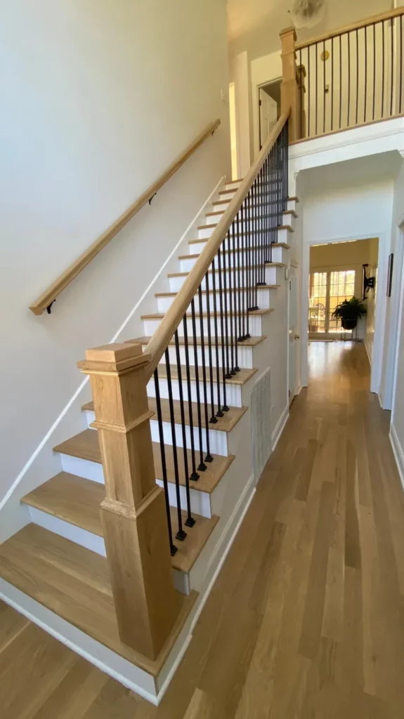 Spacious open-concept staircase featuring light oak wooden treads, white risers, square wood box newel posts, and sleek black metal iron balusters extending up to a second-floor hallway balcony.
