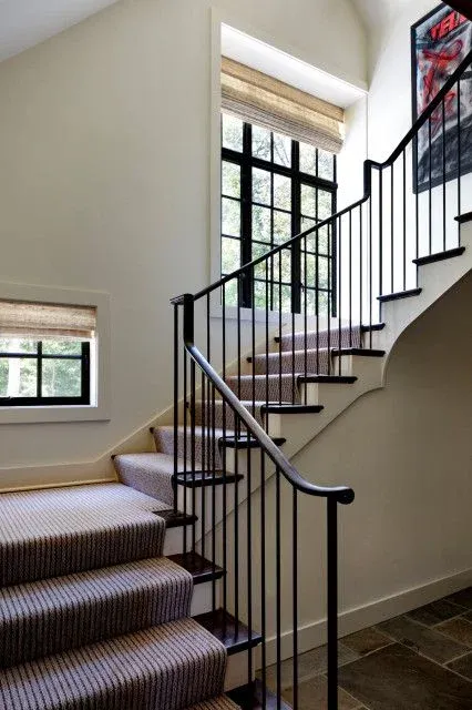 Winding staircase with dark wood treads and a textured neutral carpet runner, featuring a sleek, minimalist black iron handrail and thin vertical balusters set against off-white walls and large grid windows with woven shades.