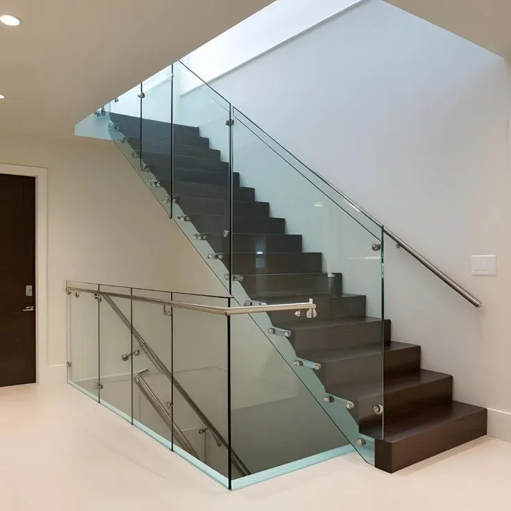 Contemporary interior staircase featuring rich dark wood floating-style treads, seamless frameless glass guardrail panels mounted with stainless steel standoff pins, and a sleek metal handrail set against clean white walls.