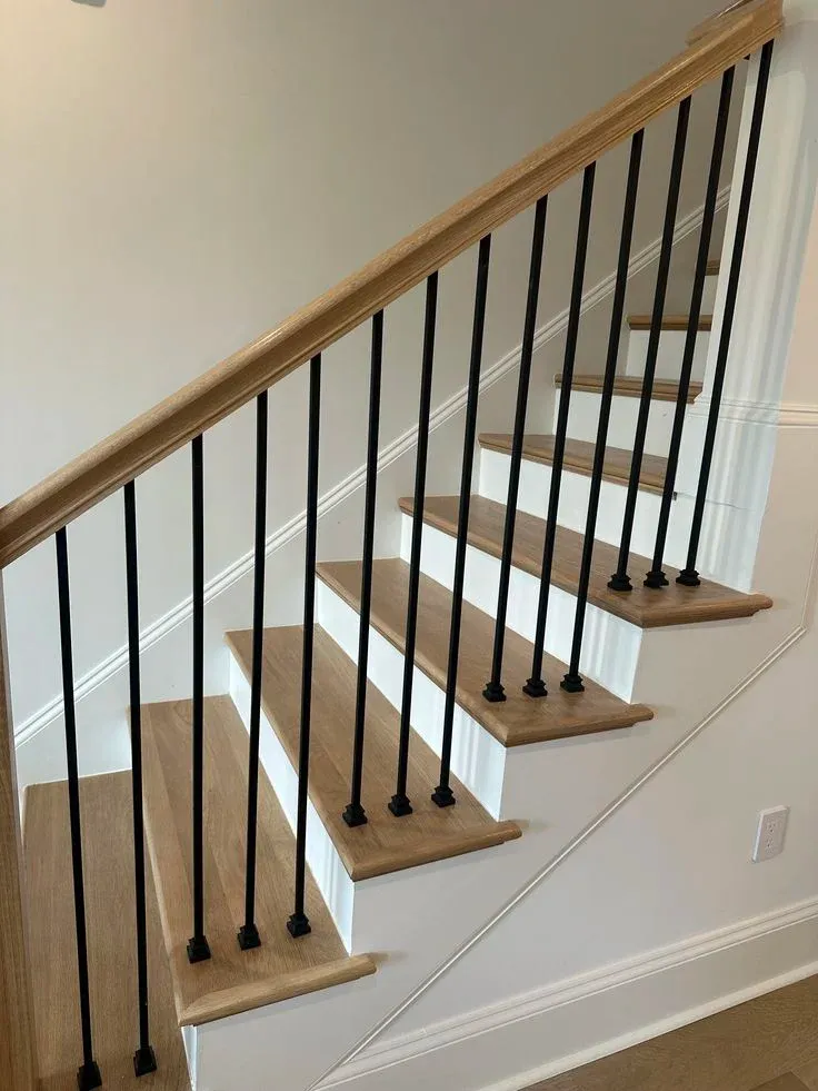 Clean contemporary staircase featuring light oak wood steps with crisp white risers, framed by a matching natural oak handrail and straight matte black metal balusters mounted with square shoes.