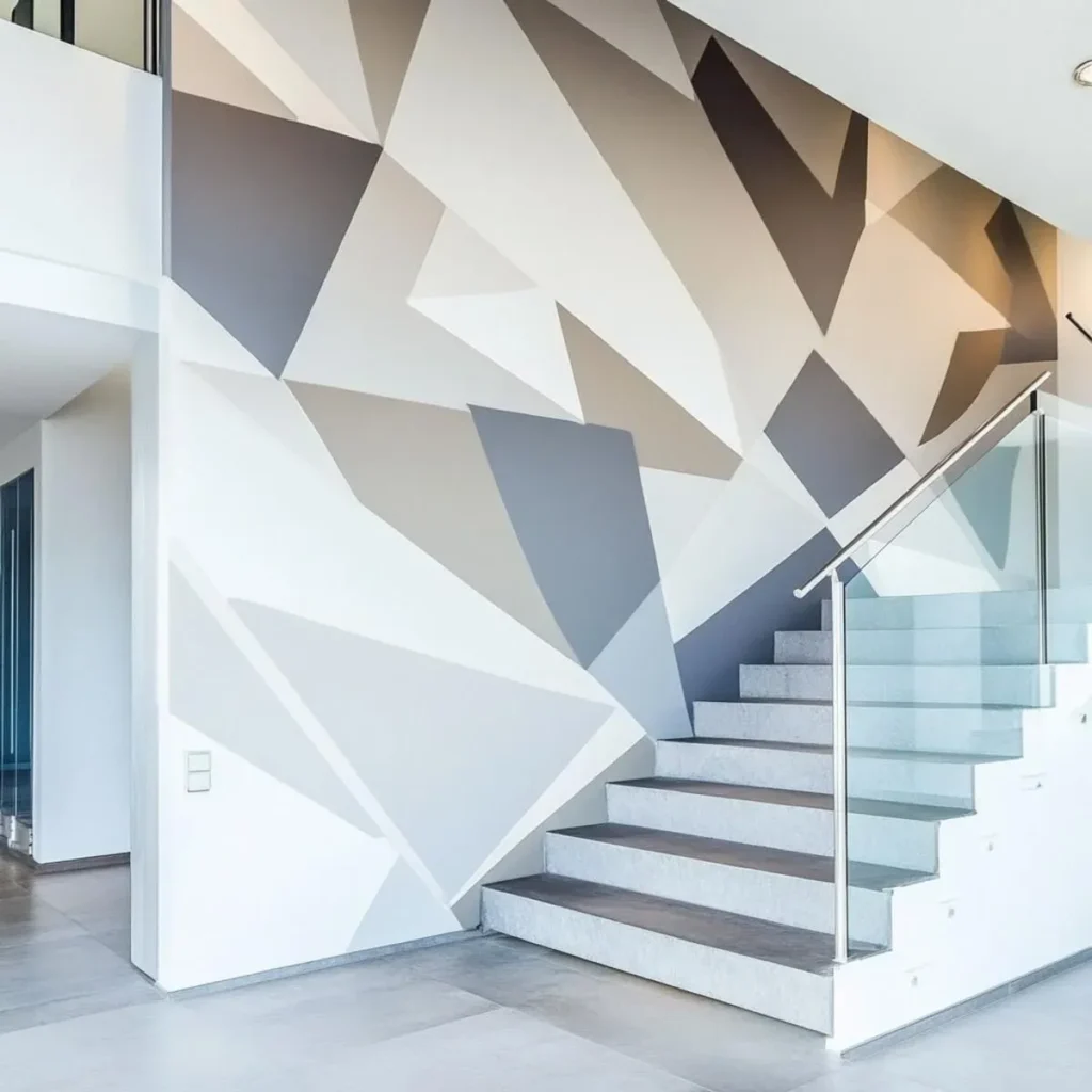 Contemporary indoor staircase featuring modern glass railings, light stone steps, and a large multi-story accent wall painted with a geometric abstract mural in neutral shades of grey, beige, charcoal, and white.