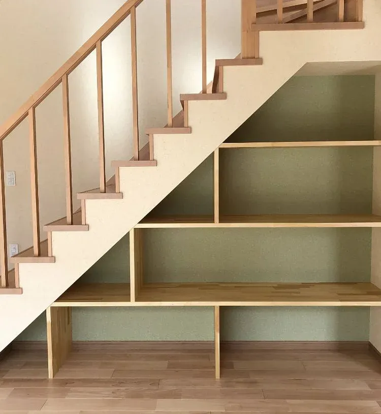 Minimalist under-stair storage area featuring custom light wood open shelving built directly beneath a matching wooden staircase with slim banisters set against a soft green accent wall and light hardwood floors.
