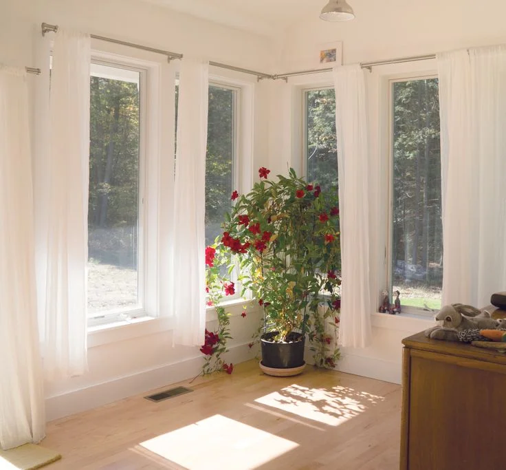 Minimalist sunroom corner design with multiple tall paneled windows, light hardwood floors, white trim, and airy white curtains letting in natural light.