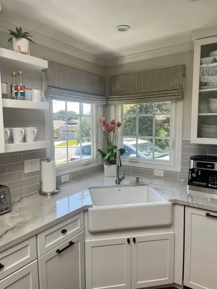 Bright kitchen corner sink setup featuring a white farmhouse apron-front sink, white shaker cabinets, grey subway tile backsplash, and two windows decorated with striped linen Roman shades.