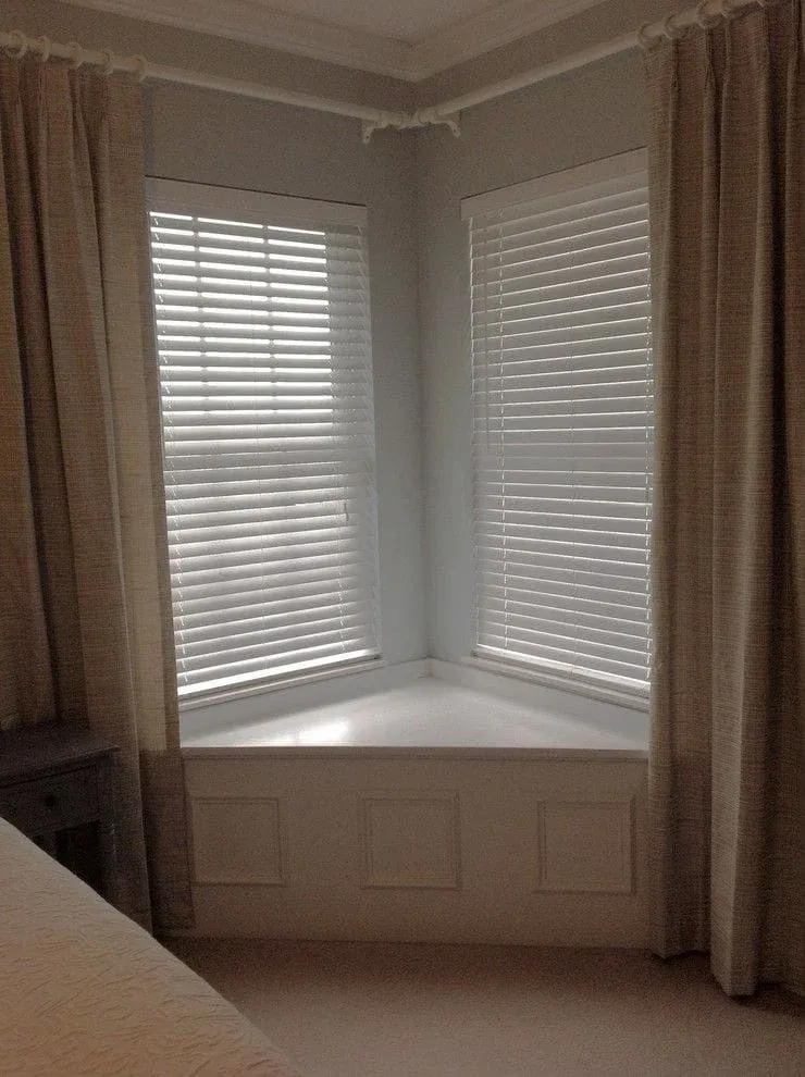 Minimalist bedroom corner design showing a white wooden triangular bench seat under dual corner windows fitted with horizontal blinds and framed by tan floor-length curtains.