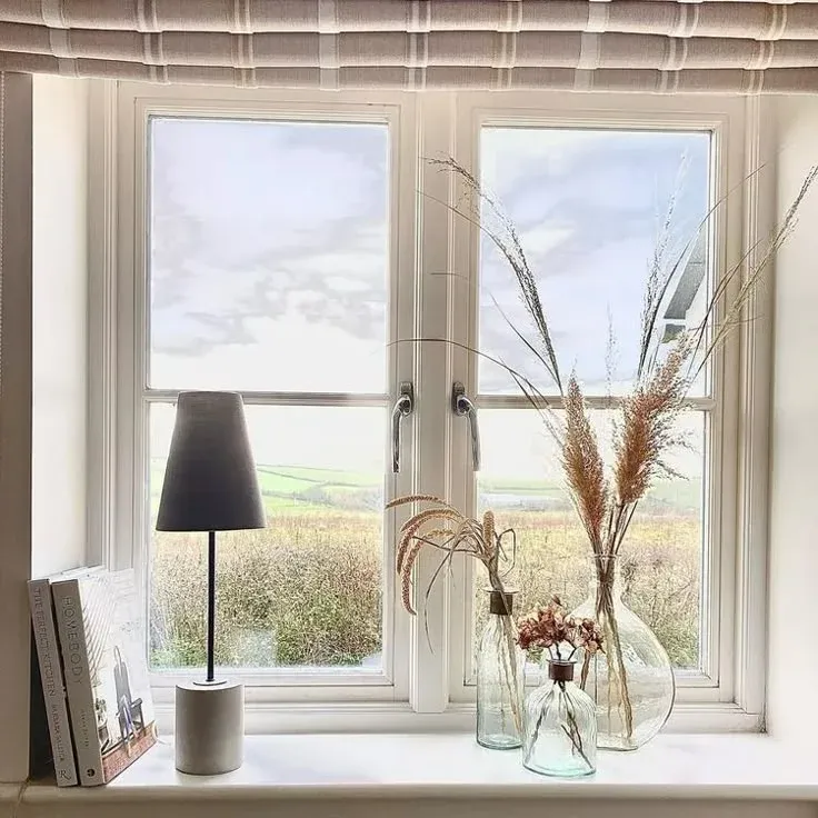 Styled deep window sill with glass bottles containing dried ornamental grasses, a small table lamp, books, and a view of the countryside.