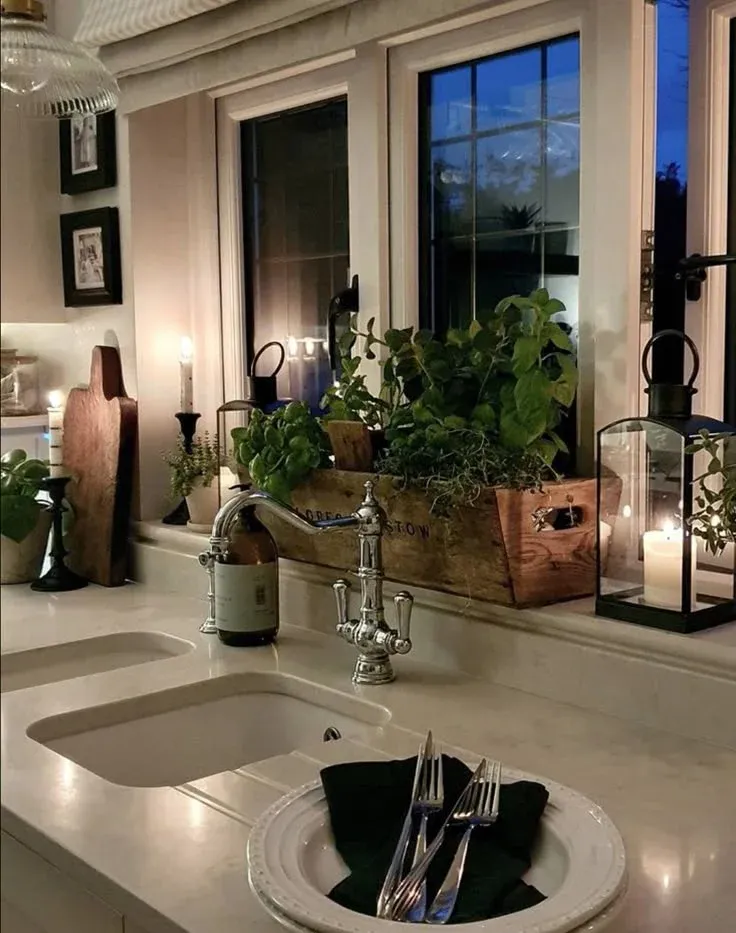 A rustic wooden planter box overflowing with fresh green herbs sitting on a deep kitchen window sill behind a classic chrome faucet, lit by glowing candle lanterns at dusk.