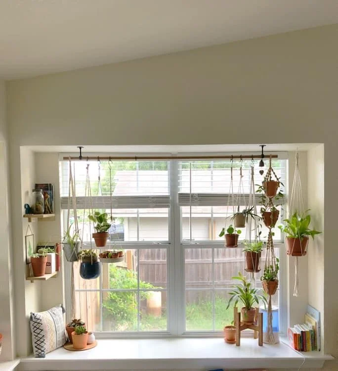 A bright window alcove decorated with a hanging plant display, small floating shelves on the side wall, a patterned throw pillow, a mini wooden plant stand, and stacks of colorful books on a wide white sill.