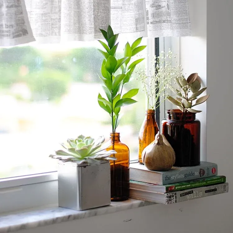 A marble window sill features a silver planter with a large succulent, alongside amber glass bottles holding dried baby's breath, green leaves, and other botanicals. A stack of decorative books supports a ceramic chicken figurine under newspaper-patterned curtains, all bathed in soft, natural sunlight.