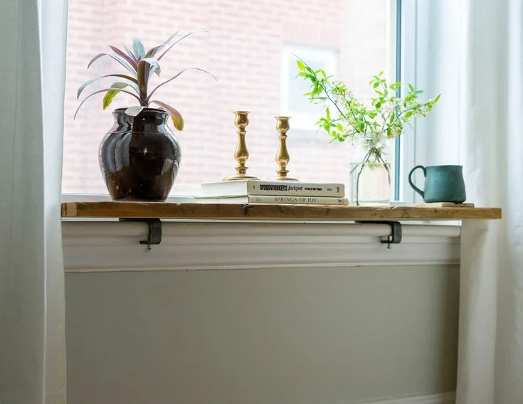 A rustic wooden shelf extension clamped securely over a white window sill, beautifully styled with a dark ceramic plant pot, stacked books, gold candlesticks, a jar of fresh green cuttings, and a teal mug.