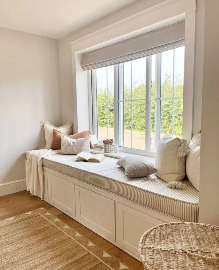 Cozy reading nook featuring a custom white built-in window bench with shaker cabinet storage, a beige and white striped cushion, and an assortment of tassel and terracotta pillows. A light grey Roman shade hangs over the large window, looking out to green trees, with a natural jute rug on the hardwood floor below.