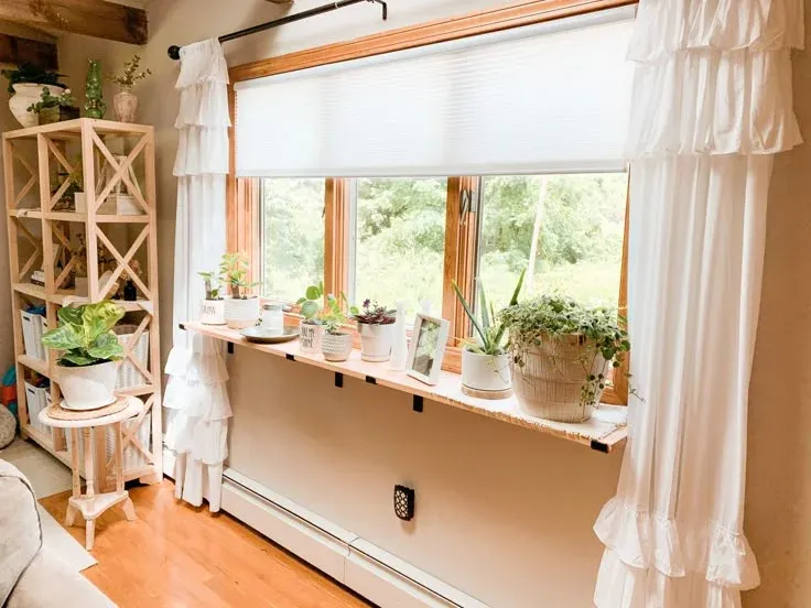 Long wooden window sill extension shelf mounted below a wood-framed window, lined with potted houseplants and picture frames.