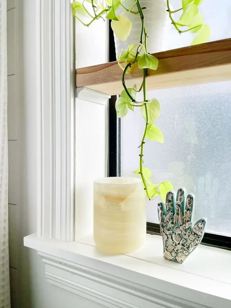 Close-up of a thick wooden shelf mounted inside a white window frame, featuring a trailing pothos plant cascading down toward a frosted glass window sill.