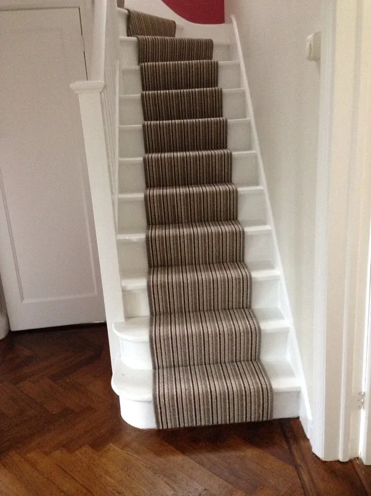 A narrow white wooden staircase fitted with a vertical brown-striped carpet runner, leading up from parquet hardwood flooring in a home hallway.