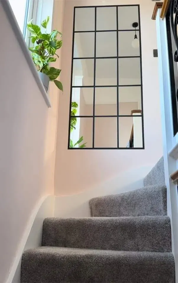 A narrow carpeted staircase featuring a large black metal grid window pane mirror mounted on a blush pink wall alongside a potted houseplant on the window sill.