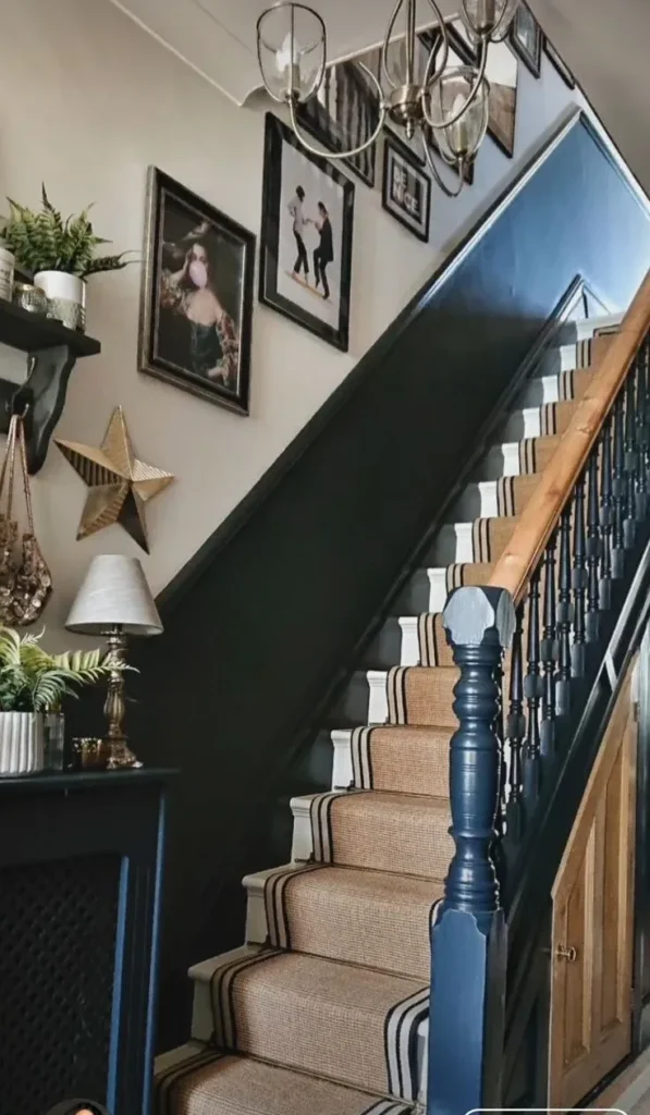 A narrow staircase featuring a dark blue painted banister, a woven jute carpet runner with black stripes, and an eclectic gallery wall with framed art prints and hanging decor.