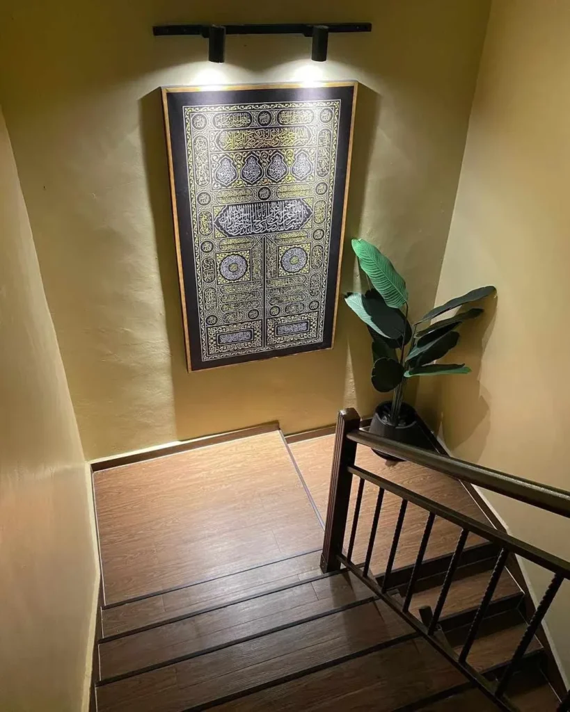 A staircase landing featuring warm lighting focused on a large framed Kiswah-style Islamic calligraphy wall tapestry, paired with a potted tropical plant on wood-look vinyl flooring.