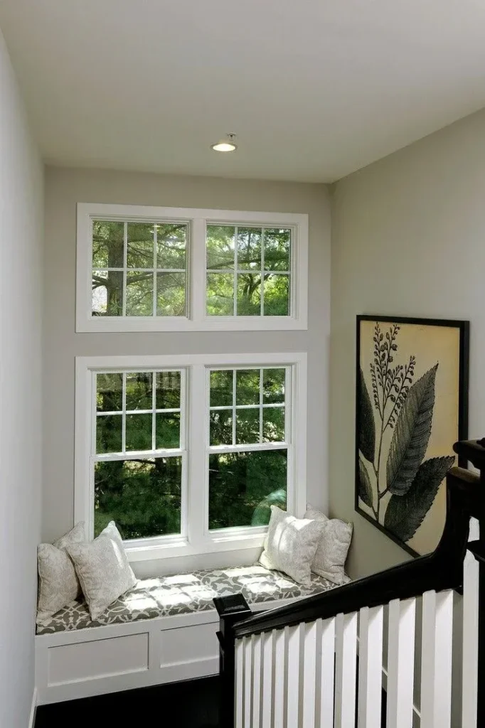 A cozy staircase landing featuring a custom built-in white window bench with patterned cushions, throw pillows, double stacked windows, and a framed botanical leaf art print.