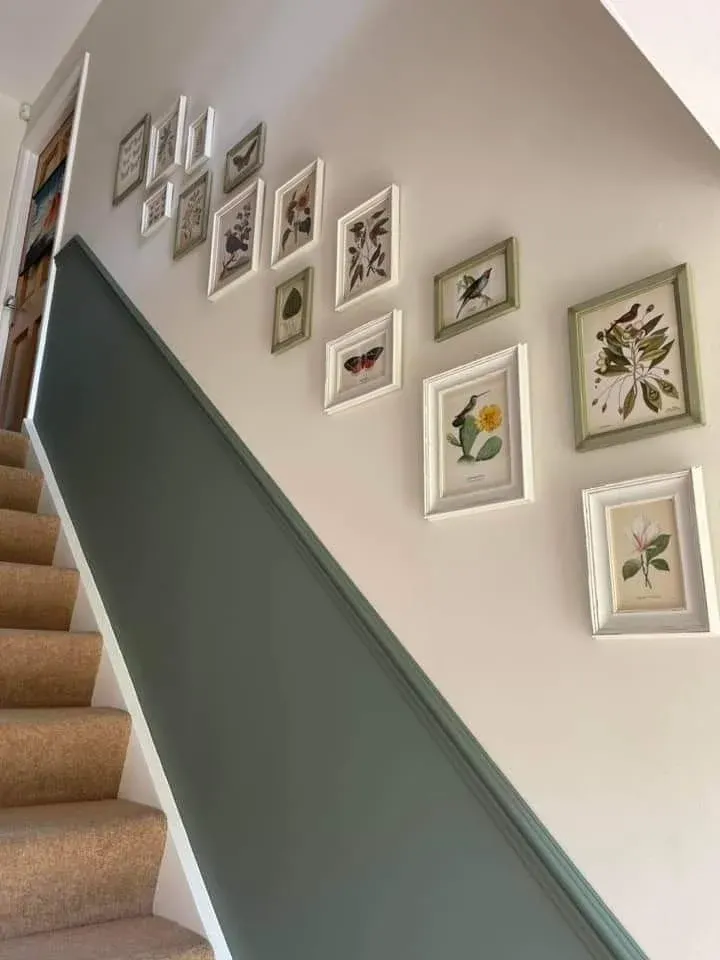 A carpeted staircase featuring a two-tone painted wall with a deep sage green lower section topped with decorative trim and a beige upper wall. A grid-style gallery wall displays small framed botanical and nature art prints in white and sage green frames following the incline of the stairs.