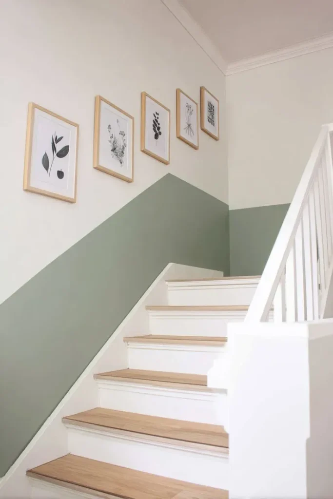 A bright wooden staircase featuring light wood treads, white risers, and a white balustrade set against a two-tone painted wall. The lower half of the wall is painted in a muted sage green at a diagonal angle following the stairs, while the upper wall is off-white with five light wood framed botanical prints aligned in a row.