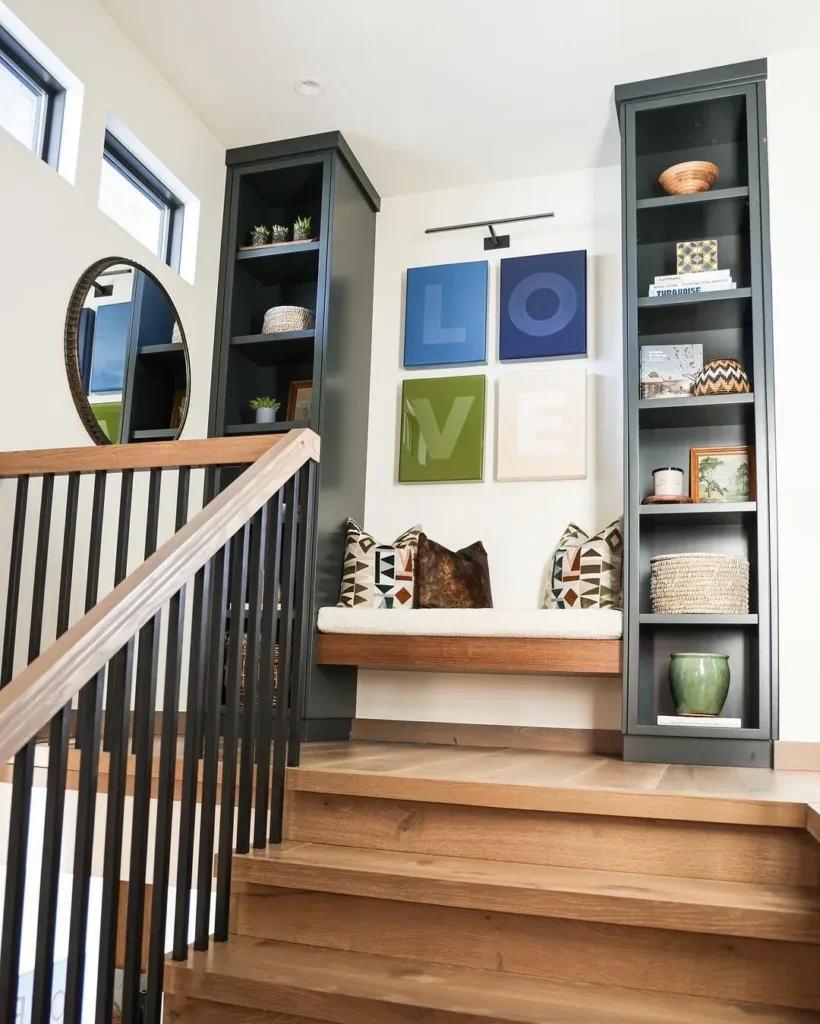 A wooden staircase landing featuring a built-in bench seat flanked by dark grey tall open bookcases, with decorative throw pillows, a four-panel modern canvas wall art spelling out "LOVE", a picture light overhead, and a round wall mirror on the left.
