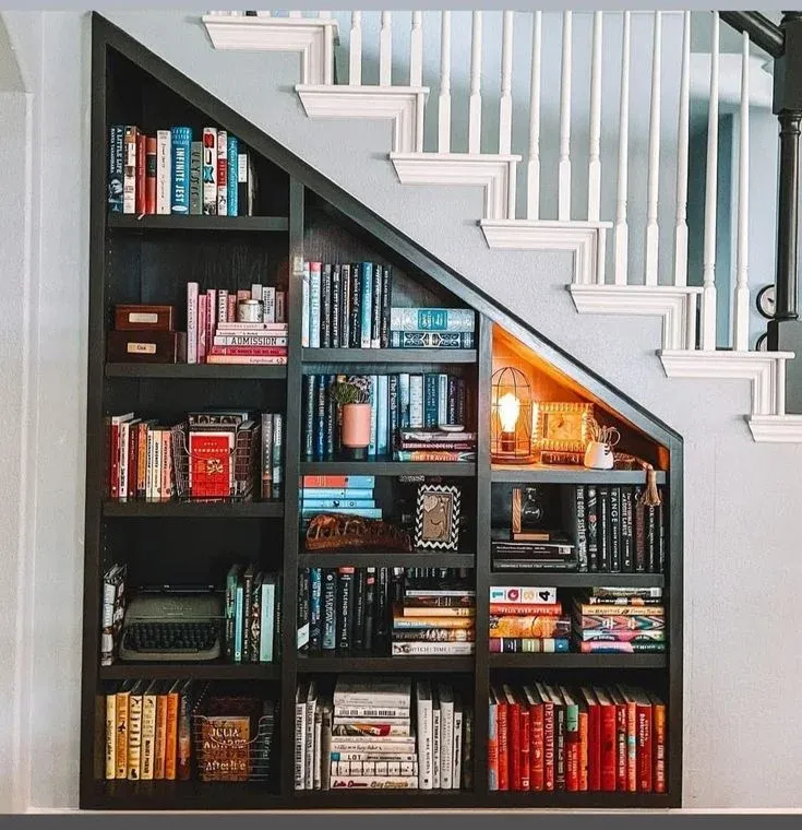 Custom black-painted under-stair bookshelf filled with colorful books, decorative items, a vintage typewriter, and a warm glowing table lamp.